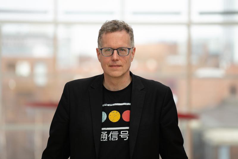 Portrett av ein mann i dagslys. Han har briller, dressjakke og ein tskjorte med kinesisk skrift på.